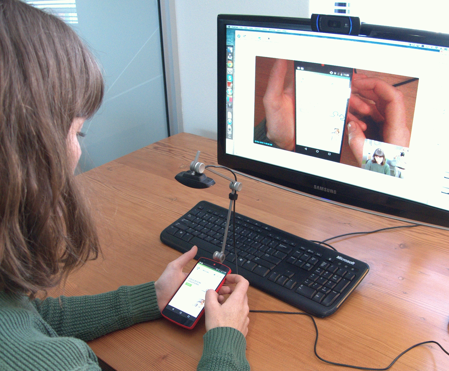 A woman conducts usability testing on a mobile phone with a camera rig attached to it which shows her phone screen on a computer monitor in the background